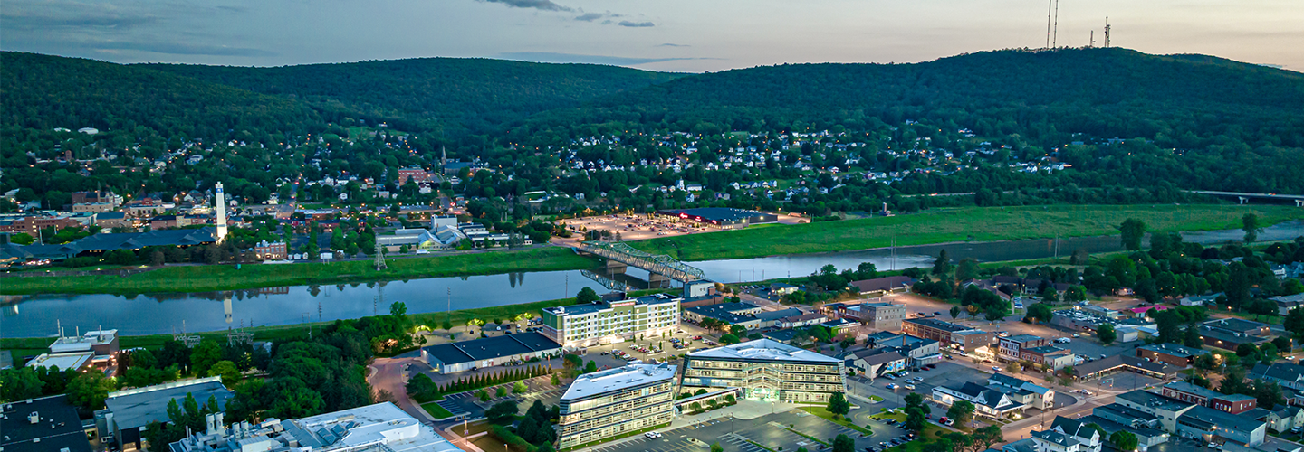 Aerial view of small city, river, bridge, and buildings; tree-covered hills with phone tower on top in the distance
