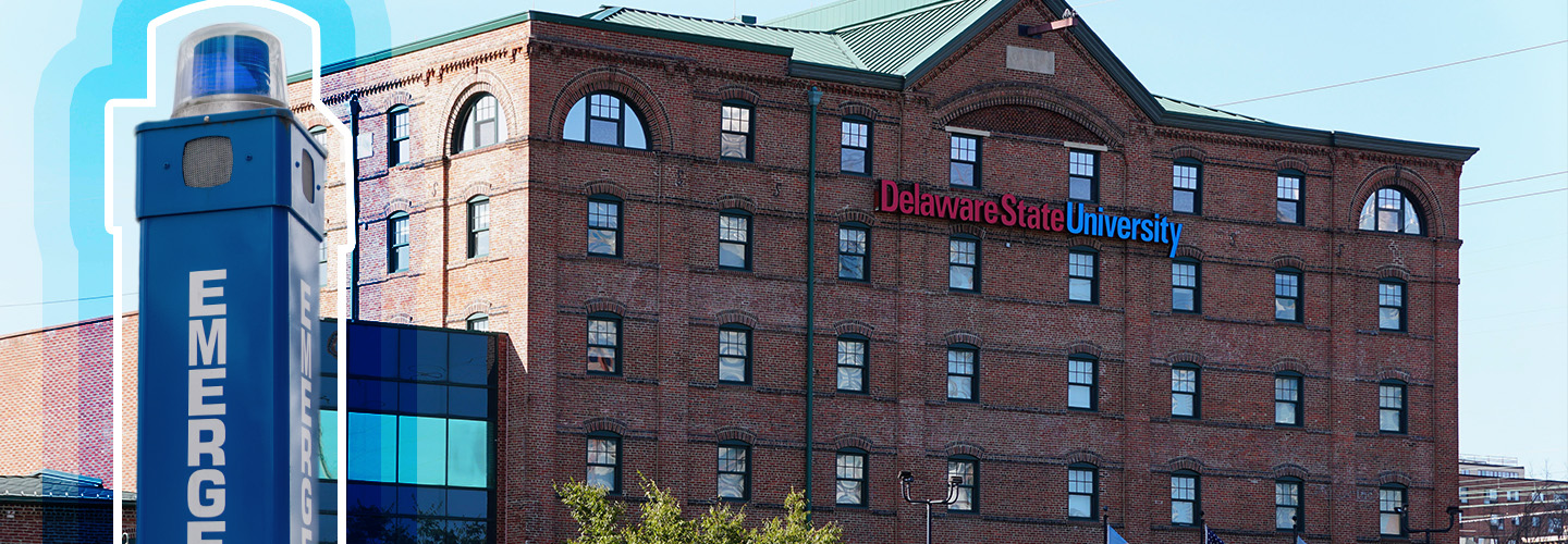 A blue light emergency phone sits to the right of a several story, campus building with Delaware State University across the facade. 