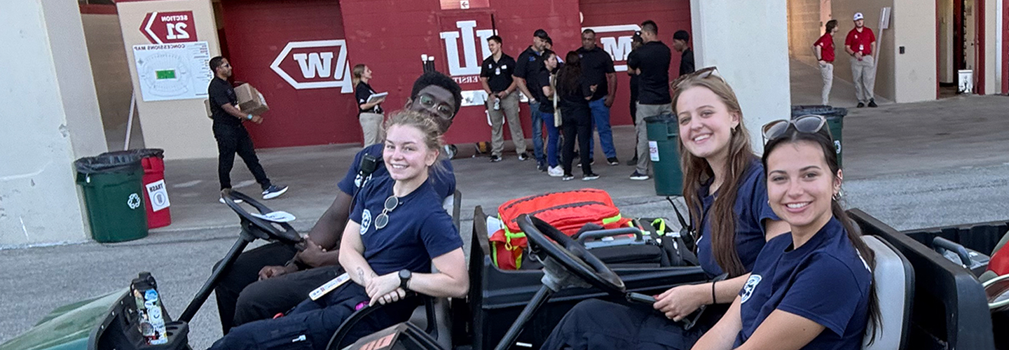 Four Intra-Collegiate Emergency Medical Service (IC-EMS) students smiling at the camera from electric carts while on duty at the Indiana University football stadium. 