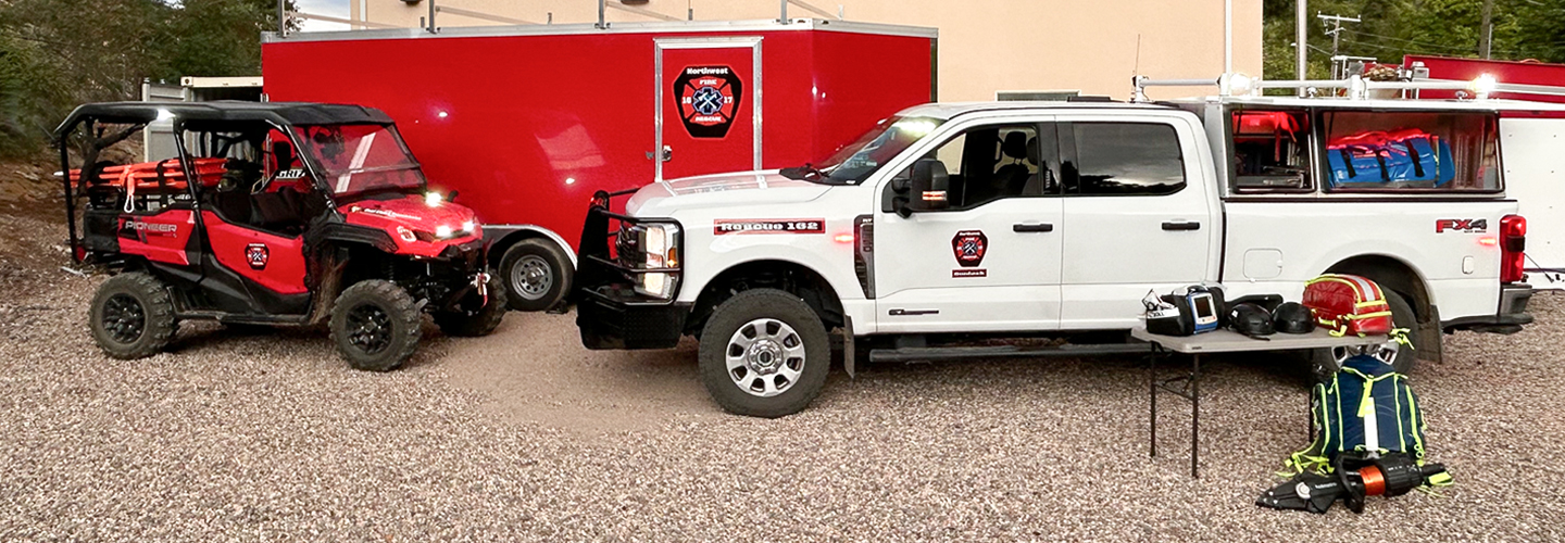 A truck, UTV, and transport trailer outfitted with FirstNet devices such as HPUE parked outside a Northwest Fire and Rescue station. 