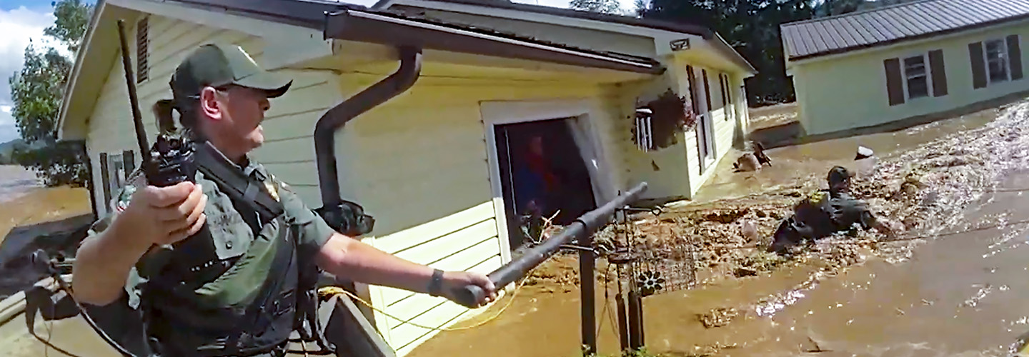 A first responder in tactical gear and holding a radio uses a pole and rope to assist a person through severe floodwaters outside a damaged home; Debris and muddy water surround the homes 