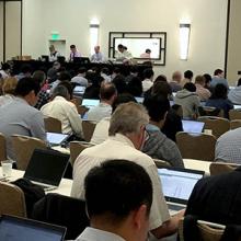 A room full of attendees at the June 2019 3GPP meetings listen and take notes on laptops as a panel of four speakers at the front give a presentation.