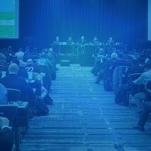 A large conference room filled with rows of people seated at long tables, facing toward five presenters sitting at a dais and two jumbo screens in front of them at a 3GPP meeting