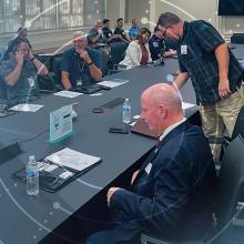 Attendees gather around a table at the FirstNet Users Summit; abstract pattern moves outward from attendees like rings on a tree 