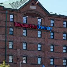 A blue light emergency phone sits to the right of a several story, campus building with Delaware State University across the facade. 