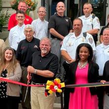 Group of officials and first responders standing in Allegany County, Maryland. Three people are holding a ceremonial ribbon, and one man cuts it with a large pair of scissors.