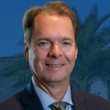 Mayor Billy Hewes’s headshot sits in front of a water tower with Gulfport written on it and foliage. 