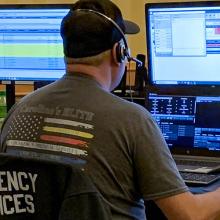 Emergency telecommunicators take calls at a desk with multiple monitors. Emergency telecommunicators take calls at a desk with multiple monitors.