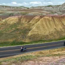 A scenic view of a winding road cutting through rocky hills in South Dakota, with a group of motorcyclists riding one behind the other along the curve of the road, surrounded by hills and rural terrain under a partly cloudy sky.