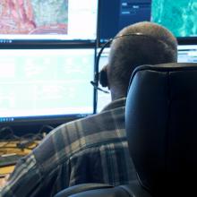 911 call-taker wearing a headset sitting at a desk in a communications center; multiple computer monitors showing maps, data, and emergency calls