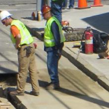 Workers in safety vests and hard hats observe a hole in the street.