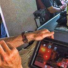 A firefighter riding in a helicopter speaks into a headset while looking down at a tablet with information
