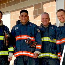 Judges for the Haptic Interfaces for Public Safety Challenge wearing firefighter personal protective equipment pose together; judge wearing firefighter gear tests haptic prototype