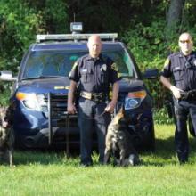 Three police officers standing in front of three Hazelton Police Department vehicles, each holding the leash of a German shepherd dog. 