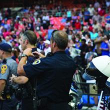Three police officers using devices stand in front of a large group of people in a stadium
