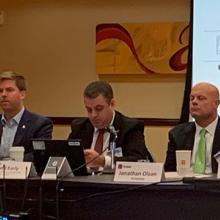 Members of the FirstNet Authority and the Public Safety Advisory Committee sit at table with slides projected behind; “December 2019 PSAC Meeting”