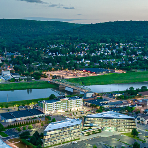 Aerial view of small city, river, bridge, and buildings; tree-covered hills with phone tower on top in the distance