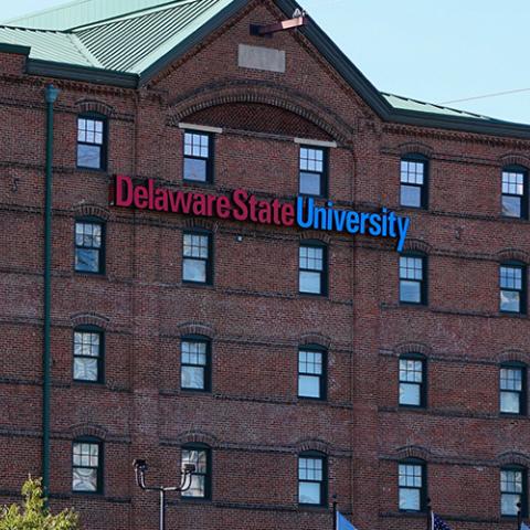 A blue light emergency phone sits to the right of a several story, campus building with Delaware State University across the facade. 