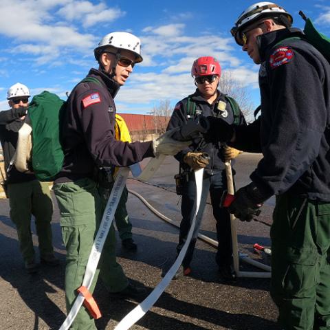 Boulder responders talk while holding a fire hose in a parking lot; City of Boulder staff member wheels out a FirstNet Compact Rapid Deployable