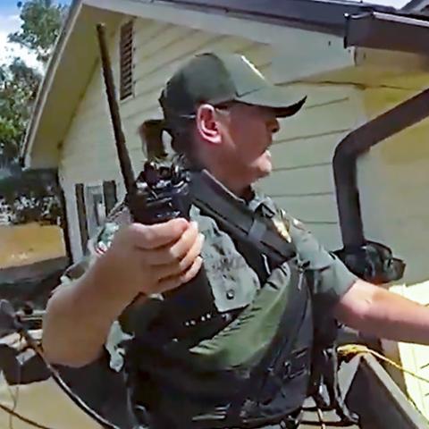 A first responder in tactical gear and holding a radio uses a pole and rope to assist a person through severe floodwaters outside a damaged home; Debris and muddy water surround the homes 
