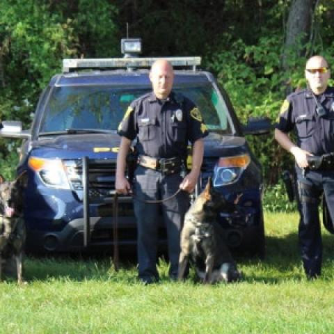 Three police officers standing in front of three Hazelton Police Department vehicles, each holding the leash of a German shepherd dog. 