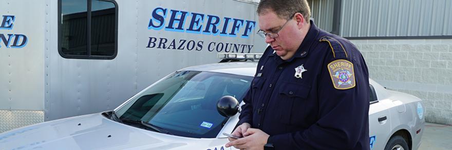 Deputy from Brazos Sheriff's Department looks at a smartphone standing in front of a mobile command vehicle