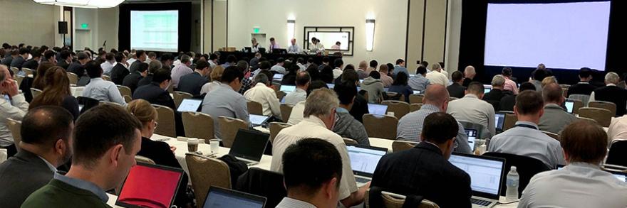 A room full of attendees at the June 2019 3GPP meetings listen and take notes on laptops as a panel of four speakers at the front give a presentation.