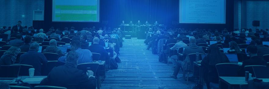 A large conference room filled with rows of people seated at long tables, facing toward five presenters sitting at a dais and two jumbo screens in front of them at a 3GPP meeting