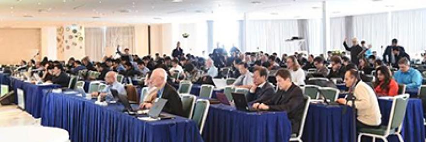 People sit at tables in a crowded room, working on their laptops and listening to a presenter.