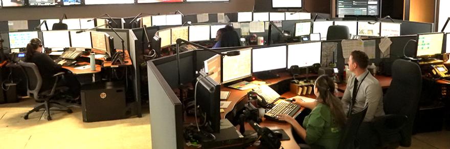 Miami-Dade dispatchers take calls and discuss issues inside of their center surrounded by information on various screens 