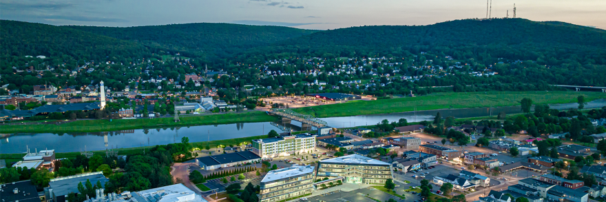 Aerial view of small city, river, bridge, and buildings; tree-covered hills with phone tower on top in the distance