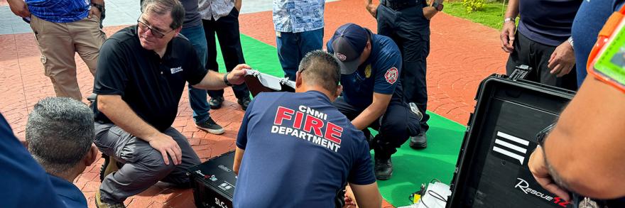 Members of the FirstNet Authority team and the Commonwealth of the Northern Mariana Islands Fire Department kneeling on the ground to demonstrate FirstNet assets during a multi-jurisdictional incident response communications exercise.