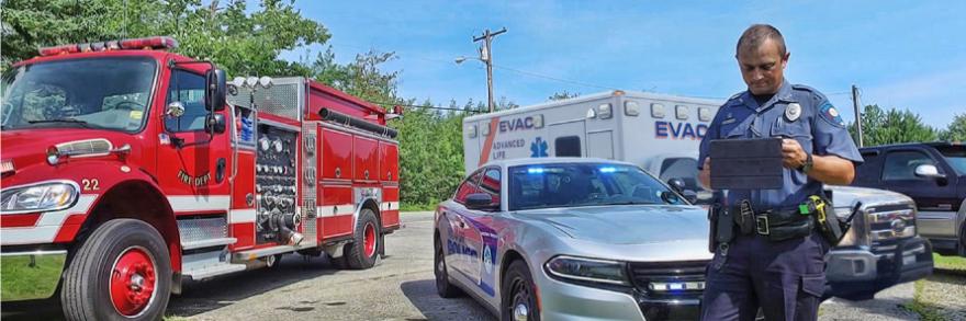 A police officer using a tablet stands in front of a fire truck, police car and ambulance.