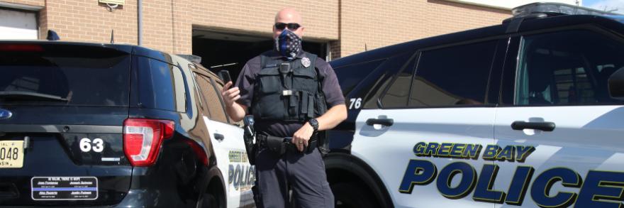 A police officer wearing a mask and a vest stands in front of a Green Bay Police vehicle