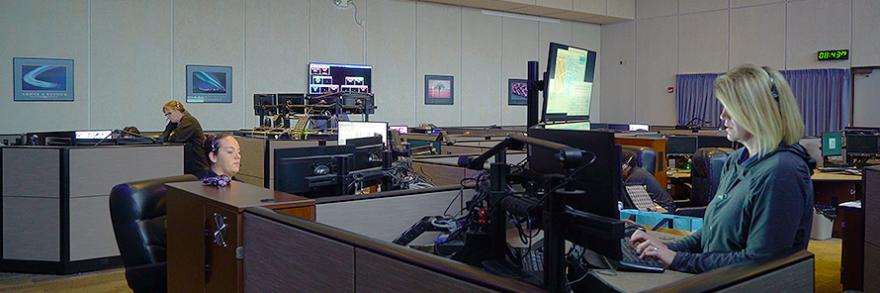 Three 9-1-1 call takers sit at dispatching consoles in a large 9-1-1 call center.