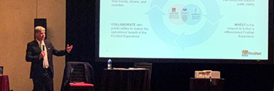 A man in a business suit stands in front of a presentation screen with his arm outstretched. 