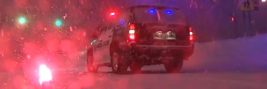 A police SUV with lights on and lit flare sit in a snowstorm at night.