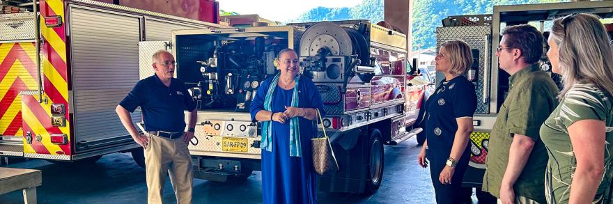 The FirstNet Authority’s Alexandra Fernández Navarro and Michael Varney and others talk with an official from the U.S. Pacific territories, standing at the rear of three public safety vehicles in an open apparatus bay.  