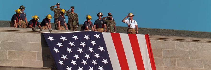 A group of firefighters in helmets and military personnel in fatigues, some saluting, standing on the roof of the Pentagon in Arlington, Virginia, unfurling a large American flag over the scarred stone.