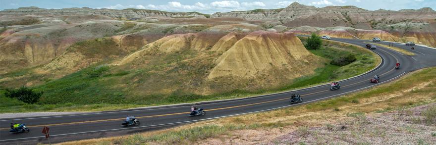 A scenic view of a winding road cutting through rocky hills in South Dakota, with a group of motorcyclists riding one behind the other along the curve of the road, surrounded by hills and rural terrain under a partly cloudy sky.