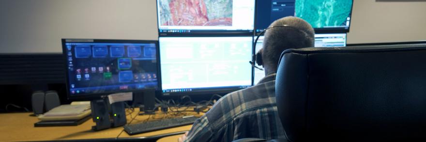 911 call-taker wearing a headset sitting at a desk in a communications center; multiple computer monitors showing maps, data, and emergency calls