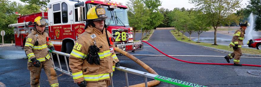 Two firefighters from the Toms River Fire Department in New Jersey carry a ladder while walking on a street in front of a fire truck 