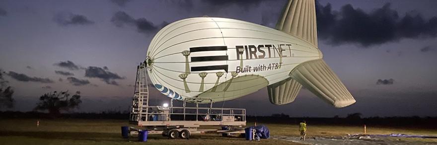 An illuminated, large aerostat, or blimp, with the FirstNet Built with AT&T logo is being prepared to take flight at night in an open field.