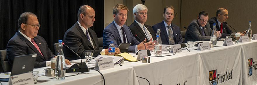 Seven of the FirstNet Authority's Board members sit at tables during a meeting in front of microphones