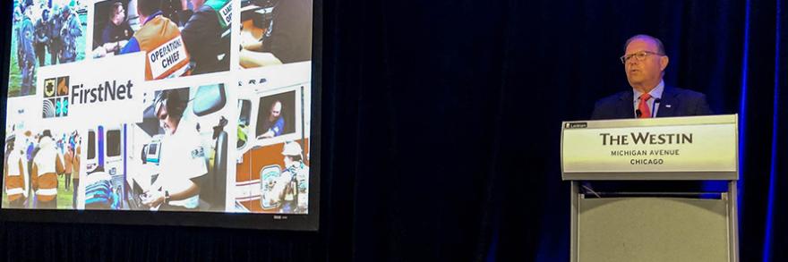FirstNet Authority Board Chair Edward Horowitz stands behind a podium with a large screen featuring the FirstNet logo and images of first responders to his right.