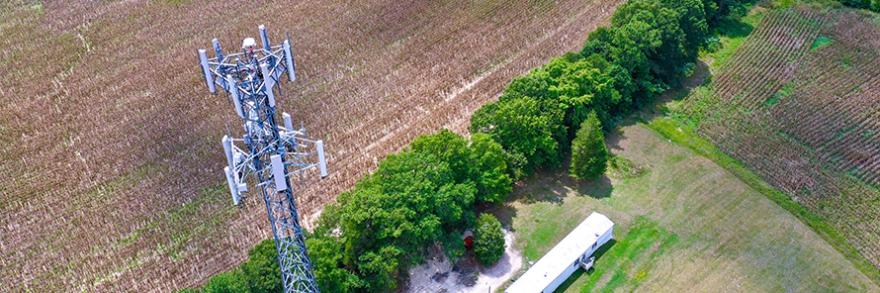 Aerial view of the top of a FirstNet cell site tower with a field and grove of trees in the background.