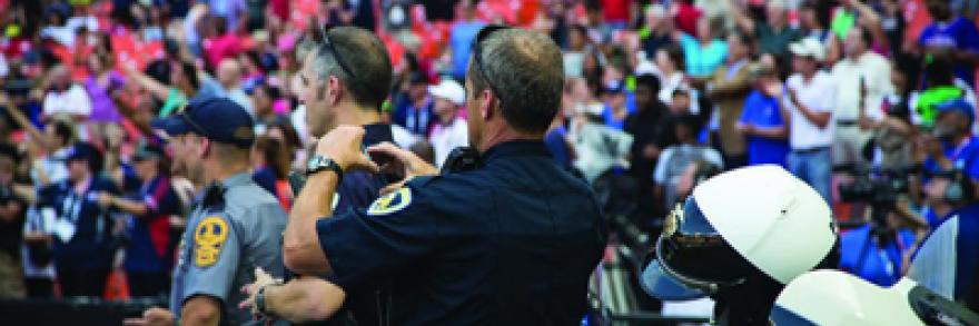 Three police officers using devices stand in front of a large group of people in a stadium