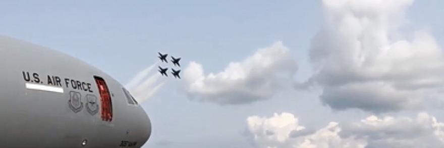 Four fighter jets streak across the sky on a summer day. In the foreground is a big crowd and a large aircraft.