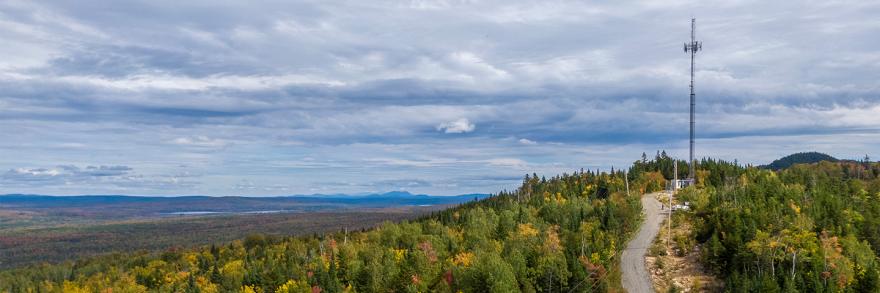 A FirstNet cell site situated on a hilly expanse surrounded by dense forest and a view of mountains and a lake in the distance.