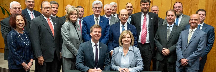 Edward Parkinson of the FirstNet Authority and LouAnn Woodward of UMMC sit in front of the Memorandum of Understanding they have just signed, behind them are standing over a dozen representatives from their organizations